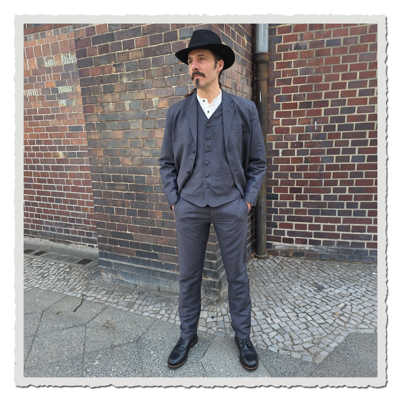 Ganzkörperansicht eines Models im grauen Nadelstreifen-Anzug vor Backsteinwand im Außenbereich.